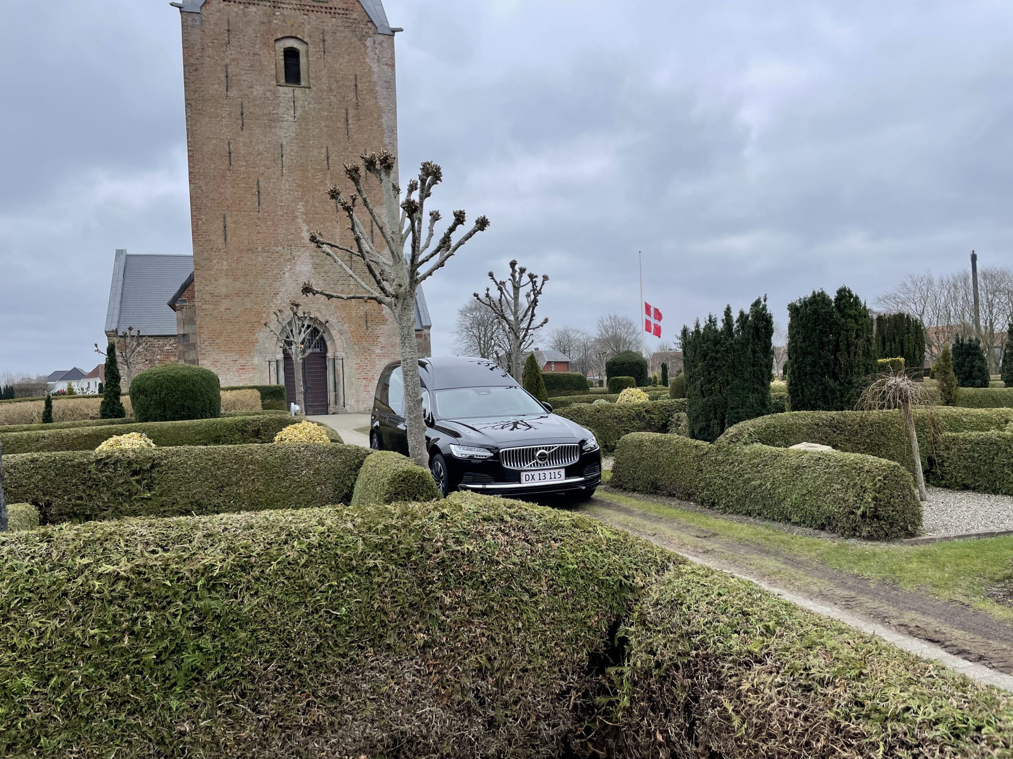 Skærbæk Kirke med en kistebil parkeret foran og dannebrog på halv stang, omgivet af veltrimmede hække under en grå, overskyet himmel.